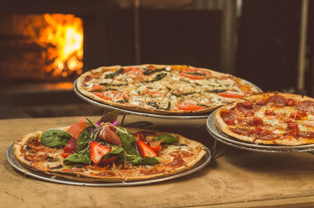 creative-approach-img Trio of gourmet pizzas with unique toppings near a wood-fired oven, ideal for food enthusiasts.